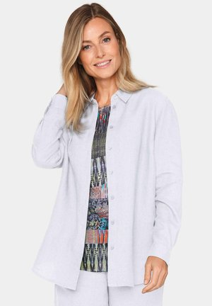 Lichtgrijze button-up shirt met lange mouwen. Zachte stof, relaxte pasvorm, kraagdetail. Hieronder is een kleurrijke, gebloemde top zichtbaar.