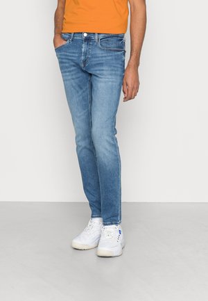 Hombre vistiendo jeans ajustados azules, camiseta naranja y zapatillas blancas, posando contra un fondo claro y liso.