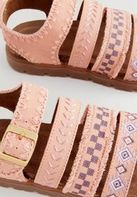 Rosa tygsandaler med fyra breda remmar, med broderade mönster och en gyllene spänne. Mjuk texturerad fotbädd och brun sula.