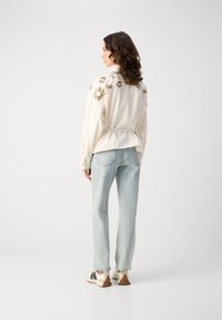 Blusa crema con ricami floreali dorati sulle spalle, abbinata a jeans blu chiaro e sneakers beige. Tessuto morbido e testurizzato, vestibilità rilassata.