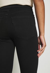 Mustat denim-housut, joissa on tiukka muotoilu, takataskut ja hienovaraiset ompeluyksityiskohdat. Kangas on sileä.