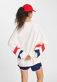 Vrouw met lang, krullend haar, gekleed in een rode pet, een witte sweatshirt met rode en marineblauwe strepen, en marineblauwe shorts, staande met haar handen gekruist achter haar rug.