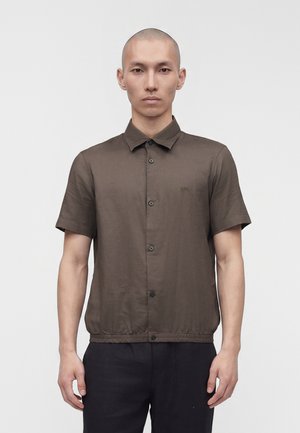 Noor mees, kes kannab pruuni lühikeste varrukatega kraega nööbitavat särki, seisab ühevärvilise heleda tausta ees.