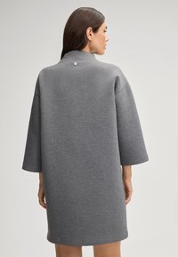 Grijze, loszittende sweatshirtjurk met driekwartmouwen, hoge kraag en een zilveren knoopdetail op de rug. Glad textuur.
