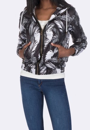 Mujer con sudadera con capucha con cremallera y estampado de hojas en blanco y negro, con las manos en los bolsillos, sobre una camisa blanca y vaqueros azules.