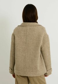 Mujer con cabello largo y oscuro, vistiendo una chaqueta de lana beige texturizada y pantalones a juego, de pie de espaldas a la cámara frente a un fondo liso.