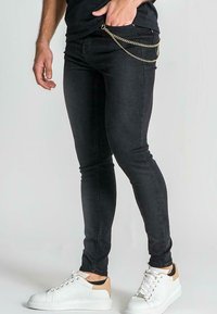 Jeans ajustados negros de denim, con un detalle de cadena en el bolsillo, combinados con zapatillas blancas con acentos en beige.