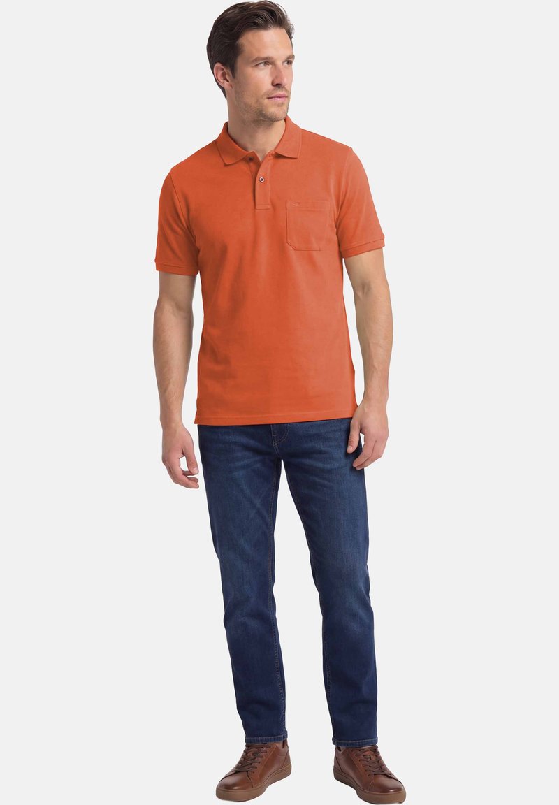 Homme portant un polo orange rouille, un jean bleu foncé et des baskets en cuir marron, debout les mains détendues le long du corps.