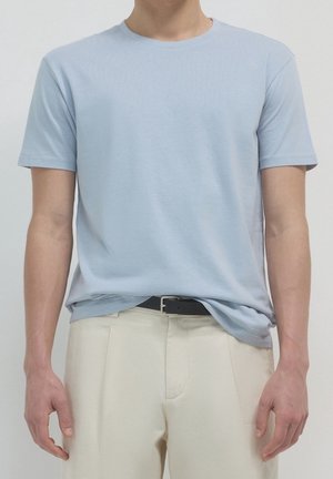 Mann trägt hellblaues T-Shirt mit kurzen Ärmeln, in cremefarbene Hosen mit schwarzem Gürtel gesteckt, gezeigt vom Hals bis zur Hüfte vor schlichter Hintergrund.