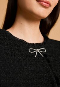 Čierna textúrovaná tkanina s rozstrapkaným výstrihom; obsahuje striebornú brošňu vo forme mašle ozdobenú jasnými kryštálmi pri zapínaní.