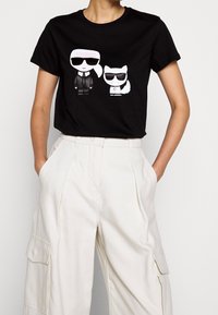 Camiseta de algodón negra con personajes de dibujos animados que llevan gafas de sol. El diseño incluye una figura en traje y un gato con una estrella.