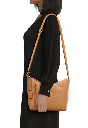 Femme en robe noire tenant un sac à main en cuir beige avec bandoulière ajustable et fermeture éclair.