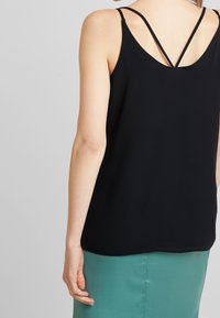 Haut sans manches noir avec des fines bretelles et un dos en V, présentant une texture lisse et une coupe décontractée. Porté avec une jupe verte.