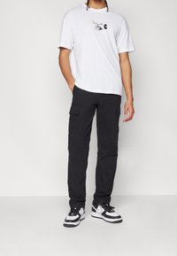 Bílé tričko s potiskem černého Mickey Mouse, doplněné černými cargo kalhotami s bočními kapsami a černo-bílými teniskami.