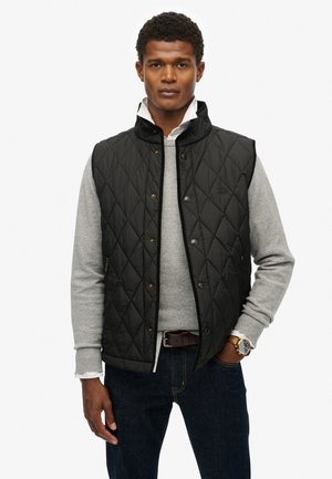 Sort quilted vest med høj krave, prydet med guld hardware og sidelommer, båret over en lysegrå sweater og mørke jeans.