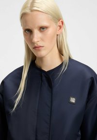Navy blue, quilted jacket with a high collar and a small metallic logo on the chest. Smooth, shiny texture with a sleek design.