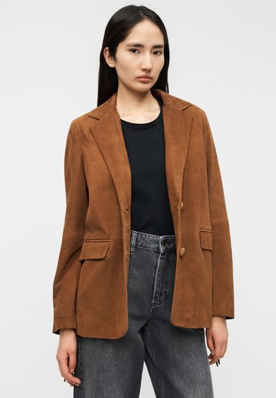 Καφέ σακάκι από σουέντ με εφαρμοστή γραμμή, με κομμένα πέτα, κλείσιμο με ένα κουμπί και δύο μπροστινές τσέπες, συνδυασμένο με μαύρο τοπ και γκρι τζιν.