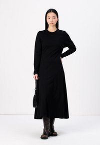 Lang svart kjole i et glatt stoff med lange ermer, slim fit og lett sveip nederst, sammen med brune skinnstøvler til knærne.