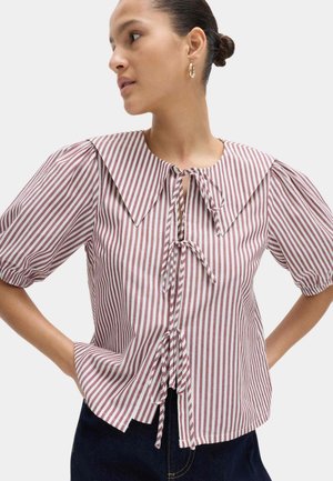 Vrouw die een blouse draagt met verticale rood-witte strepen, pofmouwen, een grote kraag en voorste striksluitingen, gecombineerd met een donkere broek.