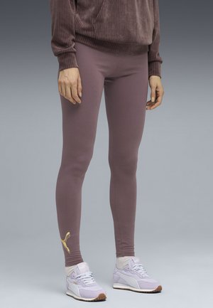 Frau, die mauve Leggings mit einem goldenen Puma-Logo am Knöchel, weißen Socken, helllila Puma-Sneakern und einem mauve gerippten Sweatshirt trägt.