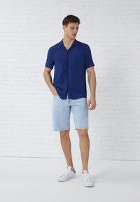 Blue short-sleeve button-up shirt paired with light blue denim shorts and white sneakers, presented against a white brick background.