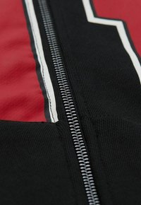 Veste zippée rouge et noire en tissu texturé avec des bandes blanches contrastantes le long de la fermeture éclair pour plus de détails.