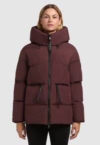 Chaqueta acolchada burdeos con cuello alto, capucha grande, cierre de cremallera y bolsillos frontales. Presenta una textura acolchada y detalles en negro.