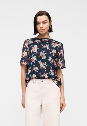 Młoda kobieta o krótkich ciemnych włosach, ubrana w granatową bluzkę w kwiaty z przezroczystymi rękawami i jasno różowe spodnie z wysokim stanem, na białym tle.