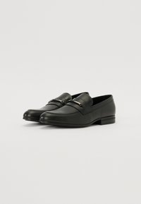 Mocasini din piele neagră cu un design elegant, având un accent metalic decorativ și un toc jos, rectangular. Textură netedă pe toată suprafața.