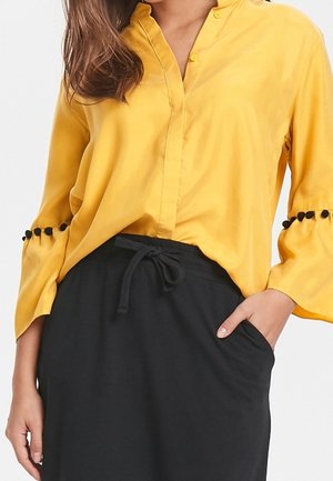 Kvinde iført en gul bluse med lange ærmer og sorte pompom-detaljer på ærmerne samt et sort nederdel med høj talje og snørebind.