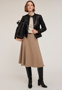 Giacca di pelle nera con colletto alto, blusa beige e gonna a tubino color beige. Stivali neri al ginocchio con texture a coccodrillo. Borsa nera in mano.