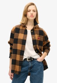 Bruin en zwart geblokt flanellen overhemd met een kraag, knoopsluiting en een borstzak. Draagt over een wit shirt en een blauwe spijkerbroek.