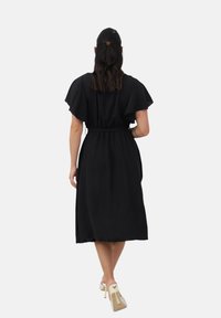 Vestido midi negro con mangas cortas tipo volante, cinturón atado en la cintura y una silueta fluida, complementado con zapatos de tacón alto transparentes.