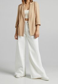 Blazer en lin beige avec manches retroussées, porté sur un crop top blanc et un jean blanc à jambes larges, associé à des baskets blanches.