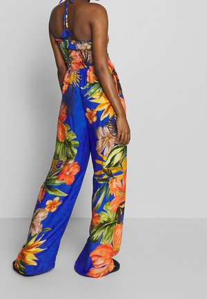 Femme debout de dos, portant un pantalon large bleu avec un imprimé floral tropical orange et vert, assorti à un haut sans manches.