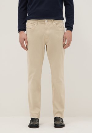 Man draagt beige broek, zwarte loafers en een donker lang mouw shirt, staat tegen een effen lichte achtergrond.