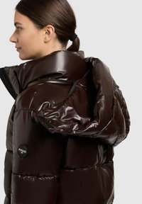 Pufferjack in glanzend, donkergroen materiaal, met een hoge kraag, ritsdetails en een zichtbaar logo-patch. Heeft een bulky, gewatteerd ontwerp.