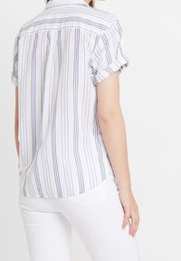 Camicia corta a righe con maniche corte, bianca e blu navy, con vestibilità comoda e maniche arrotolate. Tessuto morbido, piega sul retro per maggiore libertà di movimento.