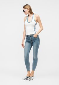 Woman wearing white tank top with blue trim, light blue skinny jeans, silver pointed shoes, and black sunglasses, standing with one hand in pocket.