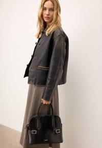 Melnas ādas jaka ar nedaudz spīdīgu tekstūru, kurai ir zelta akcenti un piecas pogas; apvienota ar melnu somu ar zelta aparatūru.