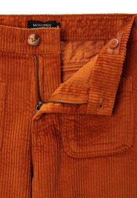 Pantalons en velours côtelé orange avec une fermeture éclair et un bouton visibles, comportant une poche avant et un tissu doux et texturé.