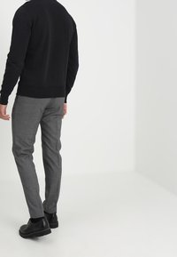Zwarte sweatshirt met ribgebreide mouwen en onderrand, gecombineerd met grijs geruite broek en zwarte schoenen, gezien van achteren tegen een witte achtergrond.
