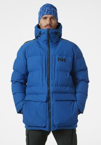 Modrá péřová bunda s vysokým límcem, zapínáním na zip, dvěma předními kapsami a černým logem. Model má také na sobě odpovídající modrou čepici.