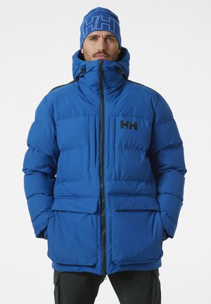 Blauwe pufferjack met een hoge kraag, ritssluiting, twee voorzakken en een zwart logo. Het model draagt ook een bijpassende blauwe muts.