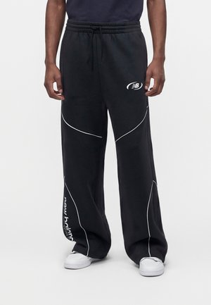 Modello che indossa pantaloni della tuta New Balance neri a gamba larga con piping bianco, logo sulla coscia, scritte vicino alla caviglia, abbinati a sneakers bianche.