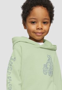 Hoodie vert clair avec une poche kangourou à l'avant, ornée d'un graphique de dinosaure et du texte "JUST BUILD" sur la manche. Texture douce.