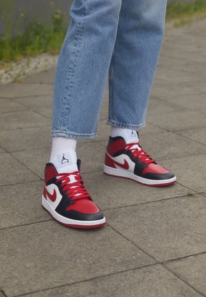 Personne portant un jean bleu, des chaussettes blanches et des baskets Nike Air Jordan rouges, noires et blanches, debout sur un trottoir gris en extérieur.