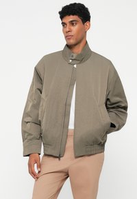 Zeleni bomber jakna s visokim ovratnikom, prednjim zatvaračem i elastičnim manžetama. Glatka tekstura i minimalistički dizajn. Nosi se preko bijele košulje.