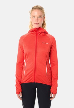 Frau mit lockigem blondem Haar, die eine rote Kapuzenjacke mit Reißverschluss und weißen Details sowie schwarze Hosen trägt, steht vor weißem Hintergrund.