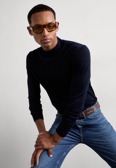 Homme portant des lunettes de soleil teintées amber, un pull col roulé bleu marine, un jean bleu et une ceinture marron, penché en avant avec les mains reposant sur un genou plié.